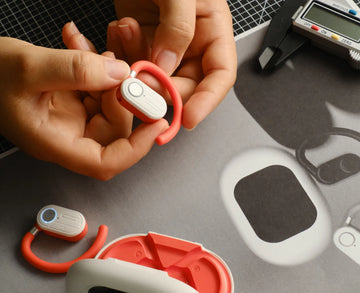 Close-up of hands holding a RELOD TouchFit PRO white wireless earbuds on a desk with electronic equipment.